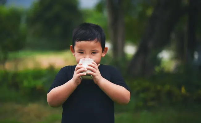 A young boy drinking a glass of milk outdoors, surrounded by greenery.