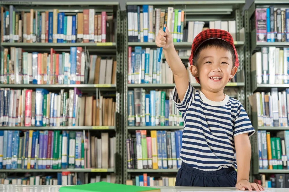 A smiling child in a striped shirt and red hat holds up a pen in a library.