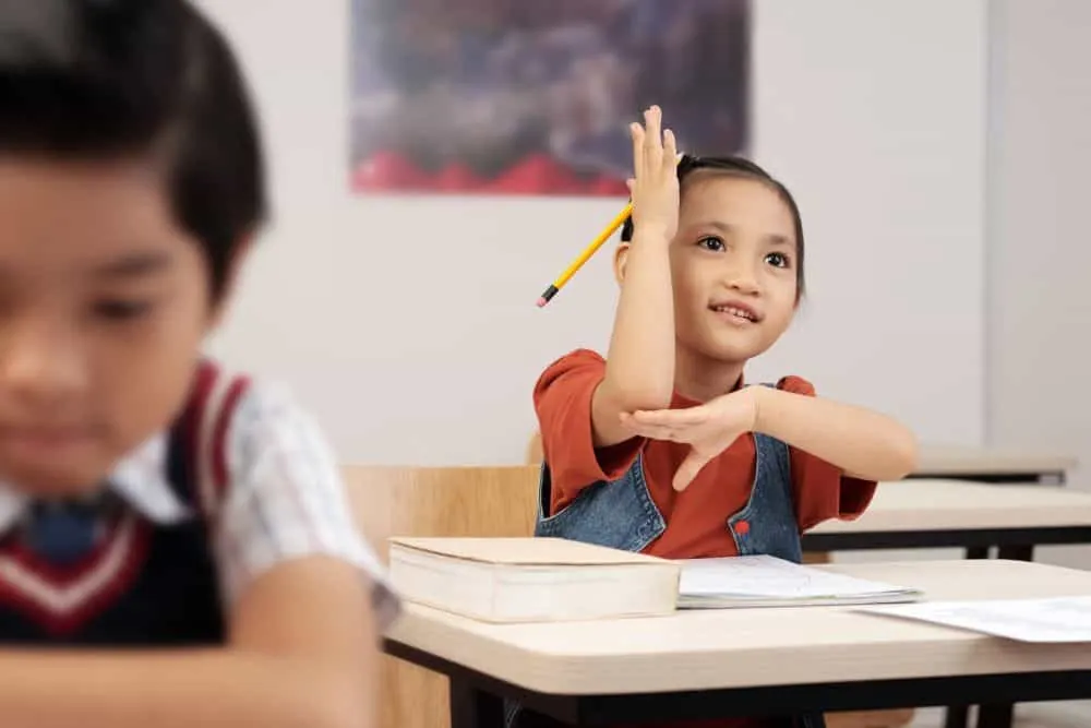A child in a classroom raises her hand, holding a pencil, with a book in front of her.